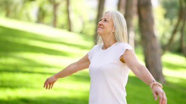 Yeşil bir parkta duran, uzun gri saçlı sakin yaşlı kadın, kollarını açıyor ve güneş ışığında gözleri kapalı derin derin nefes alıyor.