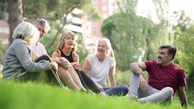 Parktaki yeşil çimlerin üzerinde oturan olgun kadın ve erkeklerden oluşan mutlu bir grup. Arkadaşlar spor yaptıktan sonra sohbet edip güneşli bir günün tadını çıkarıyorlar.