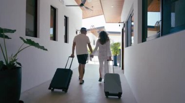 Happy young couple holding hands and walking with their wheeled suitcases through a hotel hallway. Tourists arriving at a tropical vacation resort