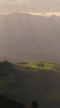 Yeşil tepeler ve ormanlarla kaplı güzel bir dağ manzarası. Altın saat güneş ışığı sabah sisinin içinden dramatik ışıklar saçıyor. Dikey görüntüler