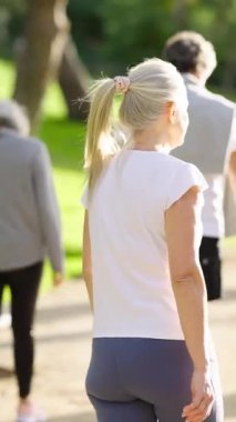 Halka açık bir parkta yürüyen olgun sarışın bir kadın. Güneşli bir günde yaşlılar için sağlıklı, aktif bir yaşam tarzı ve uygun bir ortam. Dikey