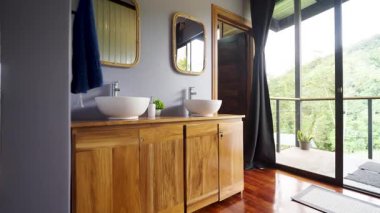 Ahşap bir dolabı, iki beyaz damar lavabosu ve aynaları olan modern banyo tasarımı. Büyük cam kapılar orman manzaralı bir balkona açılıyor.