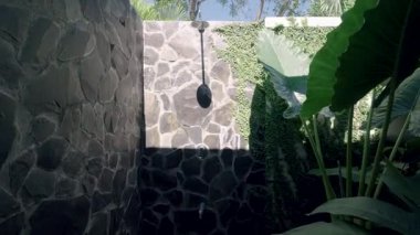 Güneş ışığı, tropikal bir tatil köyünde taştan bir duvarla dışarıda duş aldığını gösteriyor. Yemyeşil sarmaşık ve orman yaprakları tüm bölgeyi sarmış durumda.