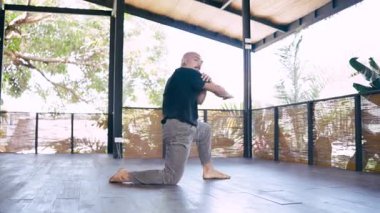 Bıyıklı kel bir adam ahşap zemini ve doğa manzarası olan güzel bir stüdyoda duygusal ve doğaçlama bir dans sergiliyor.