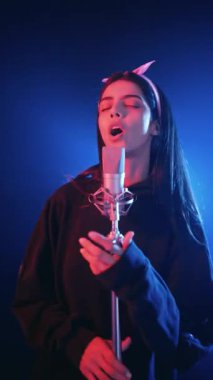 Genç bir kadın profesyonel bir mikrofona tutkuyla şarkı söylüyor. Bir kadın sanatçı canlı neon ışıkları altında yeni bir şarkı kaydediyor.