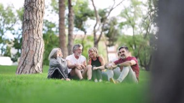 Mutlu bir grup olgun erkek ve kadın parkta çimlerin üzerinde oturup sohbet ederken keyifli bir anı paylaşıyorlar.