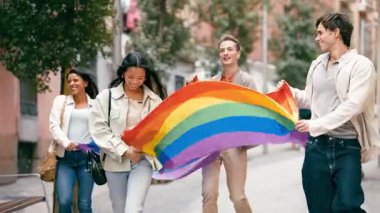 Bir grup neşeli genç arkadaş bir şehir caddesinde gururla yürürken büyük, dalgalı bir gökkuşağı bayrağı taşıyorlar.