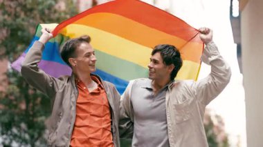 Gülümseyen genç eşcinsel çift gururu birlikte kutluyor. Gökkuşağı bayrağı tutan iki mutlu adam, aşkı, özgürlüğü ve eşitliği simgeliyor.