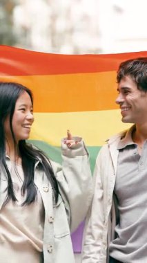 Gökkuşağı bayrağı tutan mutlu çok ırklı çift, bir geçit töreninde birbirlerine bakıp gülümsüyorlar, aşkı ve eşitliği kutluyorlar. Dikey görüntüler