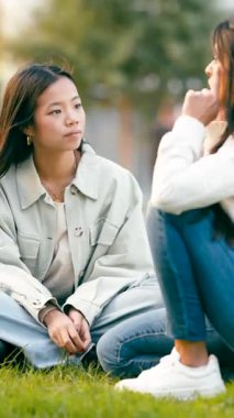 Genç Asyalı bir kadın çimlerin üzerinde oturmuş güzel güneşli bir parkta rahat bir sohbet sırasında arkadaşını dikkatle dinliyor. Dikey görüntüler