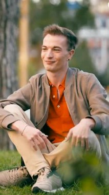 Genç yakışıklı adam çimlerin üzerinde oturuyor, gülümsüyor ve konuşuyor. Güneşli ve huzurlu bir günün tadını çıkaran bir insanın gündelik yaşam tarzı. Dikey görüntüler