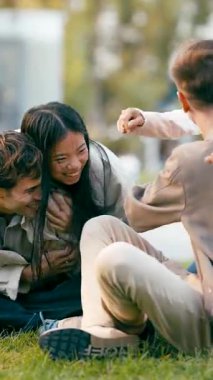 Çeşitli gülümseyen genç arkadaşlar parktaki yeşil çimlerin üzerinde otururken mutlu bir şekilde konuşuyor, sarılıyor ve birlikte gülüyorlar. Dikey görüntüler