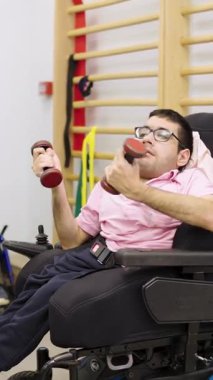 Fiziksel engelli genç bir adam tekerlekli sandalyede oturuyor ve dambıllarla çalışıyor. Rehabilitasyon egzersizleri yapan odaklanmış bir hasta.