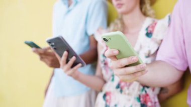 Sarı bir duvara karşı yan yana duran genç bir grup insan, her biri kendi akıllı telefonuna odaklanmış, kaydırıyor ve yazıyor.