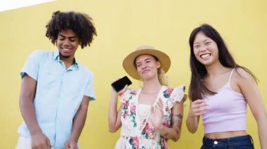 Sarı arka planda dans eden ve gülümseyen mutlu çok kültürlü genç arkadaşlar. Neşeli insanlar müzikten hoşlanıyor ve birlikte eğleniyorlar.