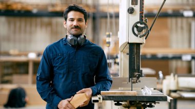 Erkek marangoz elinde bir odun parçası, profesyonel bir marangozluk atölyesinde testerenin yanında duruyor.