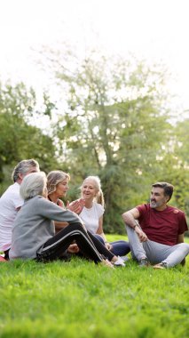 Farklı kıdemli arkadaşlardan oluşan bir grup çimlerin üzerinde otururken rahatlıyor, konuşuyor ve gülüyor, doğada birlikte olmanın tadını çıkarıyorlar.