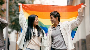 Sırıtan ve elinde gökkuşağı bayrağı tutan genç bir çift, LGBTQ artı gurur, çeşitlilik ve eşit hakların savunucusu