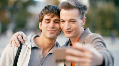 İki gülümseyen adam, eşcinsel bir çift, kucaklaşıyorlar ve akıllı bir telefonla selfie çekiyorlar, aşk ilişkilerini kutluyorlar.