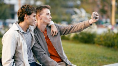 Parktaki bankta oynaşan bir selfie için poz veren genç eşcinsel çift, açık havada modern bir romantizm anını paylaşıyor.