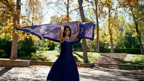 Hintli bir kadın tezahürat yapıyor, sonbaharda parkta mavi dupatta tutuyor. Neşeli bayan dans ediyor ve dışarıda güzel bir günün tadını çıkarıyor.