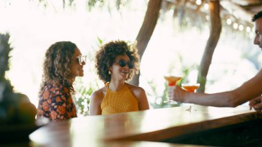 Barda dinlenen iki gülümseyen kadın arkadaş tropikal bir ortamda barmenin verdiği ferahlatıcı kokteylleri içiyorlar.