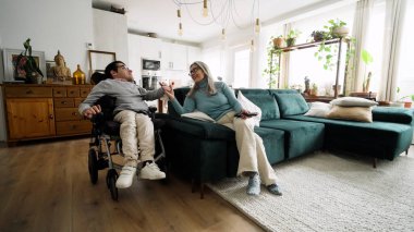 Yetişkin bir çocuk tekerlekli sandalye kullanıyor ve annesi el ele tutuşuyor. Evde sıcak bir anı paylaşıyor. Ailesinin desteğini ve duygusal bağını gösteriyor.