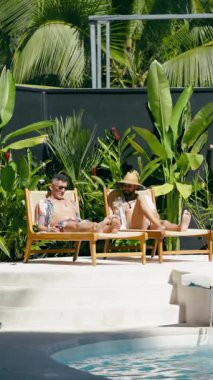 İki rahat adam havuzun kenarındaki sandalyelerde uzanıyor, konuşuyor ve yemyeşil bitkilerle dolu lüks bir tropikal otelde tatilin tadını çıkarıyorlar. Dikey