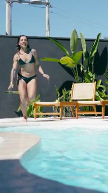 Güneşli bir günde bikinili, dövmeli neşeli genç bir kadın eğleniyor. Neşeli bir şekilde koşar ve lüks bir yüzme havuzuna atlar. Dikey