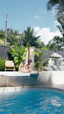 Çoklu etnik çeşitlilikte bir çiftin güneşli bir günün tadını çıkarırken, küçük bir şelalesi olan lüks bir yüzme havuzunun kenarında otururken geniş bir çekim. Dikey