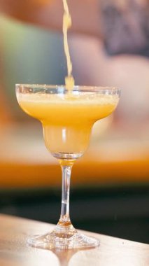 Bir barmenin tuzlu margarita bardağına canlı bir portakal kokteyli dökerken yakın plan, sonra da bir el bardan alır. Dikey