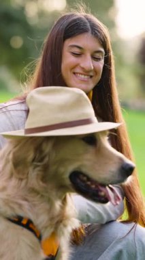 Uzun kahverengi saçlı, neşeli, bej fötr şapka takan Golden Retriever 'ıyla eğlenen mutlu bir genç kadın.