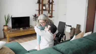 Sevgi dolu bir anne, fiziksel engelli oğlunu kanepeden kaldırır ve nazikçe tekerlekli sandalyesine yerleştirir.