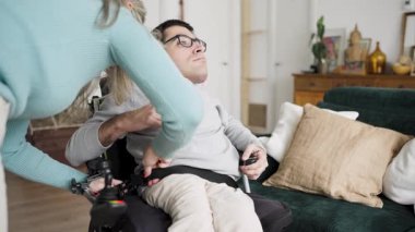 Şefkatli bakıcı, fiziksel durumu olan bir adama elektrikli sandalyesinin emniyet kemerini oturma odasına bağlayarak yardımcı oluyor.