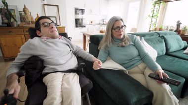 Orta yaşlı anne ve Spina Bifida 'lı yetişkin oğlu evlerinde rahat oturma odalarında televizyon izlerken el ele tutuşuyorlar.