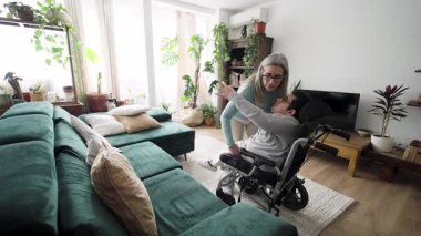 Destekleyici anne, oğlunun tekerlekli sandalyesinden oturma odasındaki kanepeye rahatlık ve dinlenme için taşınmasına yardım ediyor.