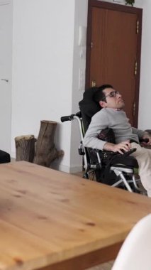 Spina Bifida ile yaşayan kararlı bir adam güçlü tekerlekli sandalyesini tahta masaya yanaştırmak için manevra yaparak evinde bağımsız hareket kabiliyetini gösteriyor.
