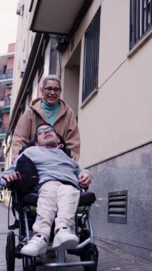 Bakıcı anne, tekerlekli sandalye kullanan yetişkin oğluyla gülüyor ve geziniyor. Mutlu aile, şehrin kaldırımında birlikte vakit geçiriyor.