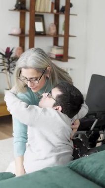Sevgi dolu bir anne, yetişkin oğluna fiziksel bir durumda yardım ediyor, onu kanepeden oturma odasındaki tekerlekli sandalyeye taşıyor.