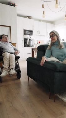 Sevgi dolu bir anne, o tekerlekli sandalyede otururken oğlunun elini okşar. Evde bakım ve bağlantı gösteren destekleyici aile anı