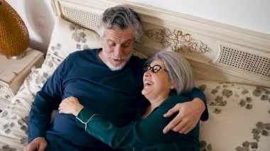 Pijamalı, yatakta yatan hoş bir yaşlı çiftin yüksek açılı görüntüsü. Erkek ve kadın gülümsüyor, sarılıyor ve sevgiyle konuşuyorlar.