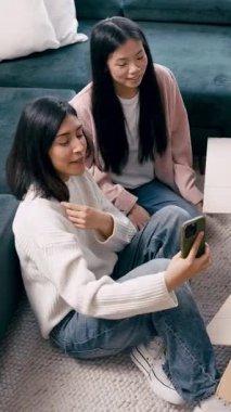 Neşeli genç lezbiyen çift gülümseyip poz verirken akıllı telefonlarıyla selfie çekiyorlar ve oturma odalarında yerde oturuyorlar. Dikey