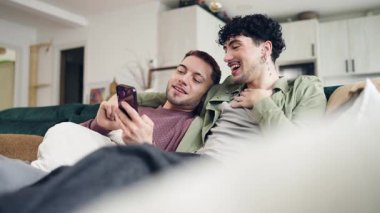 İki mutlu gay evde birlikte rahatlıyor, akıllı telefona bakıyor ve konuşuyorlar. Genç homoseksüel çift evde boş zamanlarının tadını çıkarıyor.