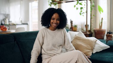Genç siyah kadın yeşil bir kanepede rahatça oturuyor, evdeki rahat oturma odasında kameraya gülümsüyor.