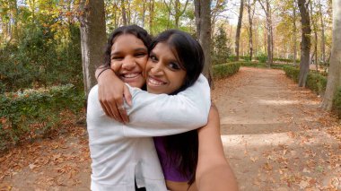 İki mutlu Hintli kadın sıkıca sarılıyor, sonbahar yapraklarıyla bir parkta kameraya gülümsüyor.