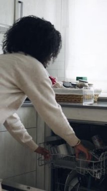 Afro-Amerikalı ev hanımı kirli bardakları ve tabakları bulaşık makinesinin rafına dikkatlice yerleştiriyor. Evde günlük ev işleri yapan genç bir kadın. Dikey