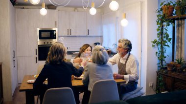 Bir grup mutlu kıdemli arkadaş bir masanın etrafında toplanıyor, bir yemek yiyor ve şarap kadehleriyle kadeh kaldırıyorlar.