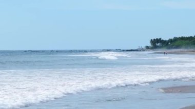 Kosta Rika 'da kumlu bir kıyıya yumuşak okyanus dalgaları vuruyor. Berrak mavi gökyüzünün altında palmiye ağaçları olan güzel tropikal bir manzara.