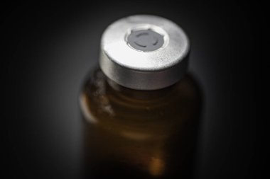 Kehribar rengi aşı enjeksiyon şişesinin seçici odak noktası olan bir makro çekim. Sade siyah zemin üzerine kurulmuş..