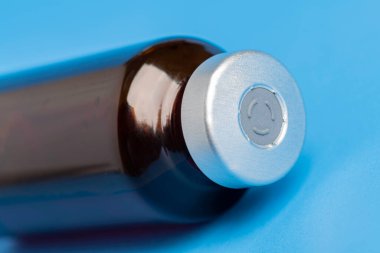 Kehribar rengi aşı enjeksiyon şişesinin seçici odak noktası olan bir makro çekim. Düz mavi arka plan üzerine kurulmuş..